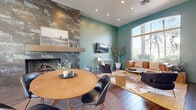 Located at 1601 E Highland Ave in Phoenix, AZ, this single-floor community lounge space offers a thoughtfully designed environment that balances comfort and entertainment. The interior is characterized by soaring ceilings, rich wood-look flooring, and a bold sage-green color palette that lends the space a modern, welcoming ambiance.

The living area serves as the centerpiece, anchored by a striking floor-to-ceiling stone tile fireplace with a sleek gold mantle. A large arched window floods the seating area with abundant natural light while offering views of the surrounding palm tree-lined streetscape. Comfortable leather seating and layered rugs create an inviting lounge atmosphere.

Adjacent to the living area, a billiards zone features a contemporary pool table with chrome legs, surrounded by additional windows that continue to bring in natural light and outdoor views. A glass sliding door connects the main lounge to a secondary space, visible through clean white walls adorned with botanical artwork, further expanding the functional footprint.

Recessed lighting throughout provides consistent illumination, complementing the natural light. The overall design blends contemporary and warm elements — from the metallic fireplace surround to the gold-accented furnishings — creating a sophisticated yet approachable communal retreat.