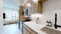 Located at 74 Freeman St in Newark, NJ, this efficiently designed one-bedroom, one-bathroom residence offers a modern living experience through clean lines, contemporary finishes, and a well-planned layout. The open-concept design maximizes the available space, allowing the kitchen, dining, and living areas to flow seamlessly together.

The kitchen is outfitted with warm wood-toned cabinetry that extends to the ceiling, stainless steel appliances including a range, over-the-range microwave, and refrigerator, complemented by white quartz countertops and a classic subway tile backsplash. Light wood-style flooring runs throughout, enhancing the bright, airy feel of the interior.

The living area is bathed in natural light from large windows dressed with sheer white curtains, offering views of the surrounding urban landscape. A dedicated dining nook with a round marble-top table adds a refined touch.

The bedroom features a calming blue accent wall, creating a distinct and restful atmosphere, while the bathroom is finished with neutral grey tile flooring and a full tub-shower combination with crisp white walls.

Decorative elements such as hanging planters in the kitchen corridor and thoughtful lighting throughout contribute to the cohesive, contemporary ambiance of this well-appointed Newark residence.