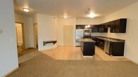 This single-floor space is designed with an open-concept layout that connects the living area and kitchen in a cohesive, functional flow. The kitchen is well-appointed with dark espresso cabinetry, granite-style countertops, and a full suite of stainless steel appliances, including a refrigerator, stove, microwave, and dishwasher. A central island provides additional counter space and serves as a natural dividing point between the kitchen and the rest of the living area. Pendant lighting hangs above the island, adding both task lighting and a modern aesthetic touch. The kitchen features wood-look hard flooring, which transitions smoothly into plush carpeting in the adjacent living space, creating a clear yet open delineation between the two zones. The living area is bright and neutral, with white walls that enhance the sense of openness. A wall-mounted shelf adds a minimalist decorative element. A door visible in the background leads to an additional room or hallway, suggesting further functional space beyond the main living and kitchen area. The overall ambiance is clean, modern, and move-in ready.