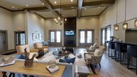 This refined communal space makes a striking first impression with its soaring coffered ceilings, detailed with dark wood beams and recessed lighting that illuminate the room with a warm, inviting glow. A dramatic floor-to-ceiling brick fireplace serves as the room's focal point, anchoring the space and adding a sense of grandeur. A mounted flat-screen television above the fireplace enhances the space's functionality for gathering and entertainment.

The lounge area is thoughtfully arranged with multiple seating groupings, including plush sofas, woven accent chairs, and upholstered armchairs, all centered around a dark round coffee table atop layered area rugs. This layout encourages both intimate conversation and larger group gatherings. A contemporary chandelier with exposed bulbs adds an industrial-chic touch that complements the space's overall aesthetic.

Natural light flows in through multiple sets of French doors with transoms, offering views to the exterior and creating a seamless connection between the indoors and outdoors. The hardwood-style flooring ties together the warm, neutral color palette that runs throughout. A secondary dining or activity area is visible in the background, further demonstrating the versatility and generous scale of this well-appointed space.