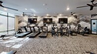 Located at 9310 Otis Ave in Lawrence, Indiana, this fitness center offers a comprehensive exercise environment thoughtfully designed for a full-body workout experience. The single-floor space is laid out to maximize functionality while maintaining an inviting, modern aesthetic.

The cardio zone features a row of treadmills and elliptical trainers positioned along a striking geometric-patterned accent wall, with multiple large flat-screen TVs mounted above for entertainment during workouts. Ceiling fans provide additional airflow throughout the room, complementing the recessed lighting overhead.

The strength training area is well-appointed with a multi-functional cable machine, a dumbbell rack stocked with a range of free weights, and an adjustable workout bench. Full-length mirrors along one wall enhance the sense of space and allow users to monitor their form. A vertical knee raise and dip station rounds out the strength equipment offerings.

Large floor-to-ceiling windows along one side of the room flood the space with natural light and provide views of the outdoors. The room also includes a practical amenity area with personal storage lockers and a water fountain, ensuring convenience for all users. Durable, patterned carpet flooring ties the entire space together, giving the fitness center a polished and professional feel.