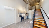 Located at 1837 Pacific Ave. in Forest Grove, Oregon, this two-story loft-style unit offers a striking industrial-modern aesthetic. The main floor welcomes you with soaring double-height ceilings, an exposed concrete ceiling with visible structural elements, and large floor-to-ceiling windows at the entrance that bathe the open living area in abundant natural light and offer views of the surrounding greenery.

The open-plan main floor features a contemporary kitchen outfitted with light wood cabinetry, stainless steel appliances including a refrigerator, electric range, and dishwasher, and a clean white countertop. A wall-mounted mini-split system provides climate control. The kitchen flows seamlessly into the living and hallway areas, enhancing the sense of openness.

A striking open-tread staircase with warm wood steps and sleek black metal railings connects the main floor to the upper mezzanine level, which overlooks the living space below through matching black metal railings. The bathroom features a wood-toned vanity with a dark countertop, an undermount sink, and a large mirror. Light wood-toned flooring runs throughout both levels, complementing the modern industrial character of the space. Pendant lighting adds warmth to the otherwise contemporary aesthetic throughout.