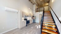Located at 1837 Pacific Ave. in Forest Grove, Oregon, this two-story loft-style unit offers a striking industrial-modern aesthetic. The main floor welcomes you with soaring double-height ceilings, an exposed concrete ceiling with visible structural elements, and large floor-to-ceiling windows at the entrance that bathe the open living area in abundant natural light and offer views of the surrounding greenery.

The open-plan main floor features a contemporary kitchen outfitted with light wood cabinetry, stainless steel appliances including a refrigerator, electric range, and dishwasher, and a clean white countertop. A wall-mounted mini-split system provides climate control. The kitchen flows seamlessly into the living and hallway areas, enhancing the sense of openness.

A striking open-tread staircase with warm wood steps and sleek black metal railings connects the main floor to the upper mezzanine level, which overlooks the living space below through matching black metal railings. The bathroom features a wood-toned vanity with a dark countertop, an undermount sink, and a large mirror. Light wood-toned flooring runs throughout both levels, complementing the modern industrial character of the space. Pendant lighting adds warmth to the otherwise contemporary aesthetic throughout.