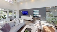 This expansive lobby makes an immediate impression with its dramatically high ceilings and an abundance of natural light streaming through large windows and glass-paneled entry doors. The herringbone-patterned hardwood flooring anchors the space with a refined, contemporary aesthetic that carries throughout the entire area.

A standout shiplap-clad fireplace wall serves as a focal point in the lounge area, rising toward the ceiling and framed by curated gallery-style artwork. Comfortable seating arrangements, including plush sofas, tufted ottomans, and accent chairs, are thoughtfully positioned around glass-topped coffee tables to create an inviting atmosphere.

A glass-enclosed reception area provides a sleek, modern check-in experience, while a dedicated workspace features a wall-mounted television, a desk, and a media console against a bold gray accent wall. A gray sliding barn door adds architectural interest and functional room division.

The lobby is further enhanced by a statement chandelier, built-in cabinetry with countertop amenities, and lush greenery throughout, lending warmth and life to the contemporary design. Overall, the space balances elegance and functionality with a bright, welcoming ambiance.