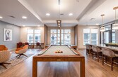 This contemporary single-floor amenity space is thoughtfully designed to serve as a multifunctional social and work hub. The open layout is anchored by a full-size billiards table set beneath a coffered ceiling with recessed lighting, creating a natural gathering point at the heart of the room. Light hardwood-style flooring flows throughout, unifying each distinct zone seamlessly.

Adjacent to the game area, a long granite-topped bar island with upholstered high-back seating is illuminated by a row of elegant gold pendant lights. Behind the bar, a textured white brick accent wall adds visual depth and dimension, complemented by built-in cabinetry and appliances that enhance the entertaining functionality of the space.

A dedicated lounge nook offers plush burnt-orange swivel chairs arranged around a round coffee table, set against a striking chevron-patterned wood accent wall. Decorative geometric screen partitions help define zones while maintaining an open, airy feel. A built-in workspace with a long wood-topped desk and seating beneath large windows provides ample natural light for those seeking a quieter corner.

Completing the space is a well-appointed coffee and beverage station, featuring a dedicated espresso machine, tea selections, a compact refrigerator, and a bold mosaic tile backsplash. Throughout, curated artwork and lush greenery add warmth and personality to this refined, amenity-rich environment.