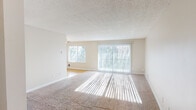 Located at 5993–6061 S 212th St in Kent, Washington, this single-floor residential unit offers a practical and comfortable layout designed to maximize livability. The open living and dining area is finished with plush wall-to-wall carpeting and neutral-toned walls, creating a bright and welcoming atmosphere. A large sliding glass door fitted with vertical blinds draws in abundant natural light and provides views of the surrounding greenery, while an additional window further enhances the airy feel of the space. A ceiling fan overhead adds both function and a finishing touch to the living area. The galley-style kitchen features white cabinetry with modern hardware, dark countertops, hardwood-style flooring, and white appliances including a refrigerator and electric range. A window at the end of the kitchen allows natural light to flow through the cooking space. The bathroom is clean and well-appointed, offering a full soaking tub and shower combination with textured surround panels and chrome fixtures. The textured ceiling finish is consistent throughout the unit, contributing to a cohesive interior aesthetic. The floor plan, as seen in the dollhouse view, reveals a thoughtfully organized layout with clearly defined living, kitchen, and private areas connected by an efficient corridor.