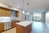 This single-floor residence showcases a thoughtfully designed open-concept layout with consistent light wood-style flooring flowing throughout. The kitchen is a standout feature, offering warm wood cabinetry, a mosaic tile backsplash, a stainless steel appliance suite including a built-in refrigerator, wall oven, gas cooktop with a sleek hood vent, and a dishwasher. A central island with a white quartz-style countertop provides additional prep space and is illuminated by pendant lighting suspended from the ceiling. The kitchen opens seamlessly into the living area, which benefits from generous natural light streaming through large floor-to-ceiling glass doors with views of surrounding trees and neighboring architecture. A ceiling fan services both the living space and additional rooms. The bedrooms are bright and well-proportioned, with large windows and light-toned walls creating a calm, neutral atmosphere. The bathroom features large-format marble-look wall tile surrounding a soaking tub with brushed nickel fixtures, complemented by a wood-toned vanity with a white countertop, an undermount sink, and a full-width mirror. Overall, the space presents a clean, contemporary aesthetic with quality finishes throughout.