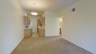 This single-story residence offers an open and functional layout that seamlessly connects the living area to the kitchen. The living space features wall-to-wall neutral-toned carpeting, creating a warm and understated atmosphere, while the kitchen transitions to tile flooring for practicality. The kitchen is equipped with light wood cabinetry that lines two walls in an efficient galley-style arrangement, providing ample storage. White appliances, including a refrigerator, stove, and microwave, complement the cabinetry. A double-basin sink is positioned along the interior wall, and a peninsula-style partial wall helps define the boundary between the kitchen and living area. A modern pendant light fixture hangs centrally above the kitchen, providing focused overhead illumination. The walls throughout are finished in a clean, neutral white, contributing to a bright and open feel. A hallway visible beyond the partial wall suggests access to additional rooms, including what appears to be a bathroom. An HVAC vent is integrated into the upper wall, and the textured ceiling adds subtle dimension to the space. Overall, the layout is practical and well-defined.