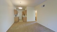 This single-story residence offers an open and functional layout that seamlessly connects the living area to the kitchen. The living space features wall-to-wall neutral-toned carpeting, creating a warm and understated atmosphere, while the kitchen transitions to tile flooring for practicality. The kitchen is equipped with light wood cabinetry that lines two walls in an efficient galley-style arrangement, providing ample storage. White appliances, including a refrigerator, stove, and microwave, complement the cabinetry. A double-basin sink is positioned along the interior wall, and a peninsula-style partial wall helps define the boundary between the kitchen and living area. A modern pendant light fixture hangs centrally above the kitchen, providing focused overhead illumination. The walls throughout are finished in a clean, neutral white, contributing to a bright and open feel. A hallway visible beyond the partial wall suggests access to additional rooms, including what appears to be a bathroom. An HVAC vent is integrated into the upper wall, and the textured ceiling adds subtle dimension to the space. Overall, the layout is practical and well-defined.