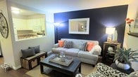 This two-floor home offers a thoughtfully designed layout that balances style and functionality. The living room serves as the heart of the main floor, immediately drawing attention with a striking navy blue accent wall that creates a bold focal point against the lighter surrounding walls. A framed mirror mounted on the accent wall adds depth and dimension to the space, while a floor lamp provides warm ambient lighting. The living area is centered around a comfortable grey sofa accented with coral and patterned throw pillows, paired with a dark wood coffee table resting on a woven area rug over hardwood-style flooring — a combination that adds texture and warmth to the room. A pass-through window seamlessly connects the living room to the kitchen, which features white cabinetry, white appliances, and a classic, clean aesthetic. This open connection between spaces promotes an airy, social atmosphere ideal for entertaining. A staircase visible through the mirror's reflection confirms access to the second floor, which expands the overall living space of the home. Together, the design choices throughout this residence create a cohesive, inviting ambiance that is both modern and welcoming.