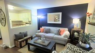 This two-floor home offers a thoughtfully designed layout that balances style and functionality. The living room serves as the heart of the main floor, immediately drawing attention with a striking navy blue accent wall that creates a bold focal point against the lighter surrounding walls. A framed mirror mounted on the accent wall adds depth and dimension to the space, while a floor lamp provides warm ambient lighting. The living area is centered around a comfortable grey sofa accented with coral and patterned throw pillows, paired with a dark wood coffee table resting on a woven area rug over hardwood-style flooring — a combination that adds texture and warmth to the room. A pass-through window seamlessly connects the living room to the kitchen, which features white cabinetry, white appliances, and a classic, clean aesthetic. This open connection between spaces promotes an airy, social atmosphere ideal for entertaining. A staircase visible through the mirror's reflection confirms access to the second floor, which expands the overall living space of the home. Together, the design choices throughout this residence create a cohesive, inviting ambiance that is both modern and welcoming.