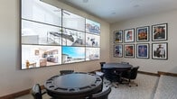 This single-floor community entertainment space is thoughtfully designed to offer a variety of social and recreational experiences. The room centers around a dedicated theater-style seating area, where plush leather recliner chairs with built-in cup holders are arranged on tiered, carpeted risers — providing an authentic cinematic viewing experience. A modern open-riser staircase with cable railings leads to an upper-level overlook area, adding architectural interest and vertical dimension to the space.

Adjacent to the theater section, the gaming area features multiple round tables, including a dedicated poker table with a padded rail and chip trays, surrounded by comfortable chairs — ideal for game nights and casual gatherings. A large multi-panel video wall dominates one wall, displaying high-resolution content across a grid of screens, serving as both a functional display and a striking focal point.

The décor throughout is cohesive and spirited, with a curated gallery of framed classic movie posters — including recognizable titles such as Top Gun, The Good, The Bad and the Ugly, Remember the Titans, and others — lining the walls and reinforcing the cinematic theme. Neutral-toned walls, patterned carpeting, and warm wood base trim complete the polished, welcoming aesthetic of this versatile community amenity space.