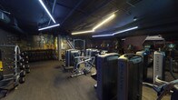 Located on Sepulveda Blvd in Sherman Oaks, Los Angeles, this fitness facility offers a comprehensive single-floor gym experience designed with a modern, industrial aesthetic. The space features an exposed dark ceiling with geometric linear LED lighting that creates a dynamic, energizing atmosphere throughout.

The main workout floor is expansively laid out, housing a robust selection of Matrix-brand strength training machines arranged for easy circulation and accessibility. A dedicated free weights area includes a full dumbbell rack and functional training equipment, complemented by a standout "Freedom Climber" climbing wall — a unique feature that adds a distinctive functional fitness element to the space.

The cardio section is well-appointed with a long row of Matrix treadmills, each equipped with individual display screens, alongside elliptical trainers and stair climbers. The dark rubberized flooring throughout provides excellent traction and durability.

A large projection screen is mounted in the training area, enhancing the workout environment. Toward the back of the facility, a convenience area includes a stocked vending machine and a lounge space with seating, offering members a place to relax before or after their sessions. Bold, mural-style artwork on the walls adds a creative urban energy to the overall ambiance.