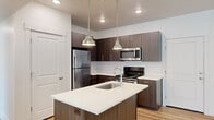 This well-designed 2-bedroom, 2-bathroom single-floor residence offers a thoughtful layout with modern finishes throughout. The open-concept kitchen and living area forms the heart of the home, where a stylish kitchen island with a white quartz countertop and an undermount sink serves as a central gathering point. Rich dark wood-grain cabinetry lines the kitchen walls alongside a full suite of stainless steel appliances, including a refrigerator, electric range, and over-the-range microwave. Pendant lighting above the island adds a refined touch, while recessed ceiling lights provide consistent illumination. Light wood-tone luxury vinyl flooring flows seamlessly through the kitchen and into the open living area, which benefits from a window that brings in natural light and maintains a clean, bright ambiance. Both bedrooms are comfortably sized, featuring the same warm flooring, crisp white walls, and windows with blinds. The primary bedroom includes a closet, while both rooms offer a calm and neutral palette. The two bathrooms are finished with dark wood-grain vanities, white quartz countertops, large mirrors, and chrome fixtures, maintaining the cohesive modern aesthetic found throughout the residence.
