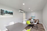 Located at 333 Holly Creek Ct in Spring, TX, this single-floor community space offers a well-organized and functional layout divided into two distinct areas. The main room serves as a versatile meeting and study space, featuring multiple gray rectangular tables paired with a mix of white and vibrant green chairs that add a pop of color to the otherwise clean, neutral palette. The walls are finished in crisp white, with a shiplap accent wall providing subtle textural contrast. Recessed lighting illuminates the space evenly, while a wall-mounted mini-split climate control unit ensures year-round comfort. Natural light filters in through windows fitted with blinds, and framed abstract artwork adorns the walls, contributing to a polished, contemporary ambiance. Wood-look flooring runs throughout, adding warmth and continuity. A second, quieter room functions as a dedicated library, furnished with open metal bookshelves stocked with a curated collection of books and accented by a geometric wall art piece. A pair of soft blue lounge chairs and a small side table create an inviting reading nook. Together, these two areas form a thoughtfully designed space well-suited for study, collaboration, and quiet reflection.