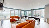 Located at 100 Aqua Way in Newport, Kentucky, this contemporary open-plan space seamlessly integrates a living room, dining area, and kitchen into a single, well-appointed floor. Large floor-to-ceiling windows flood the interior with natural light and frame impressive panoramic views of the surrounding cityscape and landscape, creating a bright and airy atmosphere throughout.

The living area is anchored by a striking brick fireplace column with a wall-mounted television, complemented by warm wood built-in shelving and cabinetry. Rich orange leather sofas arranged around a wooden coffee table add a bold, inviting contrast to the cool blue-grey accent walls and neutral large-format tile flooring that runs throughout the space.

The dining area features an expansive communal table with modern bar-height seating, ideal for group gatherings. Bold abstract artwork adorns the walls, reinforcing the modern aesthetic. Adjacent to the dining space, the kitchen is outfitted with wood-toned lower cabinetry, under-counter appliances, and additional bar-style high-top seating positioned to take full advantage of the sweeping window views. Recessed ceiling lighting throughout ensures a well-lit environment at all times. The overall ambiance is modern, sophisticated, and social.