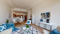 This contemporary single-floor residence showcases a thoughtfully designed open-concept layout where the living, dining, and kitchen areas flow seamlessly together. The living area is anchored by light hardwood flooring, soaring ceilings, and large floor-to-ceiling windows that flood the space with natural light while framing impressive city skyline views. A glass-top coffee table and stylish furnishings complement the airy, modern aesthetic throughout.

The kitchen is outfitted with warm wood cabinetry extending to the ceiling, crisp white countertops, a full suite of stainless steel appliances including a refrigerator, range, microwave, and dishwasher, and recessed lighting that highlights the clean, contemporary design. A peninsula counter provides additional workspace and casual seating.

The bedroom is a serene retreat, featuring plush carpet, generous windows with sweeping urban views, and an abundance of natural light that enhances the calm, neutral palette. The bathroom offers a refined finish with large-format tile flooring, floor-to-ceiling marble-tiled tub surround, a wood-panel vanity, and clean-lined fixtures that echo the modern sensibility found throughout the space. Together, these elements create a cohesive, sophisticated living environment.