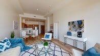 This contemporary single-floor residence showcases a thoughtfully designed open-concept layout where the living, dining, and kitchen areas flow seamlessly together. The living area is anchored by light hardwood flooring, soaring ceilings, and large floor-to-ceiling windows that flood the space with natural light while framing impressive city skyline views. A glass-top coffee table and stylish furnishings complement the airy, modern aesthetic throughout.

The kitchen is outfitted with warm wood cabinetry extending to the ceiling, crisp white countertops, a full suite of stainless steel appliances including a refrigerator, range, microwave, and dishwasher, and recessed lighting that highlights the clean, contemporary design. A peninsula counter provides additional workspace and casual seating.

The bedroom is a serene retreat, featuring plush carpet, generous windows with sweeping urban views, and an abundance of natural light that enhances the calm, neutral palette. The bathroom offers a refined finish with large-format tile flooring, floor-to-ceiling marble-tiled tub surround, a wood-panel vanity, and clean-lined fixtures that echo the modern sensibility found throughout the space. Together, these elements create a cohesive, sophisticated living environment.