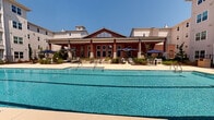Located at 9246 Highland Creek Pkwy in Charlotte, NC, this thoughtfully designed outdoor amenity area serves as a central gathering hub for the surrounding multi-story residential community. The centerpiece is a generously sized, resort-style swimming pool with crystal-clear blue water, lap lanes, and a separate wading or spa area equipped with accessible entry features. The pool deck is finished with a smooth, light-colored surface and furnished with lounge chairs and shaded umbrella seating, creating an inviting environment for relaxation under the Carolina sun. Flanking the pool is a striking red brick clubhouse building featuring soaring vaulted rooflines, large windows, and a covered outdoor patio — a standout architectural element that anchors the space. On the opposite side, a landscaped courtyard offers a peaceful retreat with winding walkways, mature magnolia trees, ornamental shrubs, and thoughtfully placed outdoor seating and grilling stations. The multi-story residential buildings surrounding the amenity area feature private balconies that overlook the pool and green spaces below, connecting indoor and outdoor living seamlessly. The overall ambiance is clean, modern, and community-oriented, designed to encourage an active and social lifestyle.