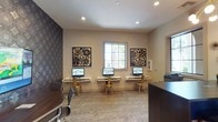 This single-floor space offers a polished and functional environment designed to serve as a business center or leasing office. The room is finished with neutral warm-toned walls and recessed lighting that provides bright, even illumination throughout. A standout feature is the row of three wall-mounted workstations, each equipped with a desktop computer monitor and accompanied by a gold metal chair, creating a sleek and organized workspace for residents or visitors. Natural light filters in through a gridded window centered on the main wall, adding a welcoming brightness to the room. Flanking the window are two boldly designed decorative art pieces featuring intricate floral motifs in rich, dark tones, which add a sophisticated and artistic character to the space. A third matching art piece is positioned to the right, maintaining a cohesive aesthetic. The flooring features a subtly patterned carpet that complements the overall refined décor. A textured dark accent wall on the left side adds depth and visual contrast to the neutral palette. A dark wood reception or administrative desk is partially visible in the foreground, reinforcing the space's professional, service-oriented function. Overall, the space balances practicality with thoughtful design details.