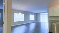 This single-floor space offers a clean, modern aesthetic defined by its open layout and thoughtful design details. The main living area is expansive and airy, bathed in natural light that streams through multiple large windows fitted with horizontal blinds, as well as a full-length vertical blind-covered sliding door or window panel that further brightens the interior. The floors throughout are finished in a sleek hardwood-style material, lending warmth and continuity to the space. A pendant light fixture hangs elegantly from the smooth, white ceiling, adding a refined focal point to the room. The walls are painted in a soft, neutral tone that complements the light-filled atmosphere and creates a calm, welcoming ambiance. White baseboards provide a crisp contrast against the flooring, reinforcing the clean lines of the interior. The open floor plan allows for flexible furniture arrangements and promotes a sense of flow throughout the living area. Overall, this space combines practical design with bright, inviting interiors, making it well-suited for a variety of uses and lifestyles.