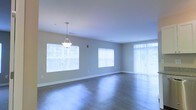 This single-floor space offers a clean, modern aesthetic defined by its open layout and thoughtful design details. The main living area is expansive and airy, bathed in natural light that streams through multiple large windows fitted with horizontal blinds, as well as a full-length vertical blind-covered sliding door or window panel that further brightens the interior. The floors throughout are finished in a sleek hardwood-style material, lending warmth and continuity to the space. A pendant light fixture hangs elegantly from the smooth, white ceiling, adding a refined focal point to the room. The walls are painted in a soft, neutral tone that complements the light-filled atmosphere and creates a calm, welcoming ambiance. White baseboards provide a crisp contrast against the flooring, reinforcing the clean lines of the interior. The open floor plan allows for flexible furniture arrangements and promotes a sense of flow throughout the living area. Overall, this space combines practical design with bright, inviting interiors, making it well-suited for a variety of uses and lifestyles.
