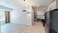 This thoughtfully designed 2-bedroom, 1-bathroom unit is laid out on a single floor, offering an efficient and comfortable living environment. Light wood-look flooring runs throughout, creating a bright, cohesive aesthetic that ties each room together seamlessly.

The open living and dining area serves as the heart of the unit, highlighted by two striking teal pendant lights that add a modern touch. Natural light filters in through a window dressed with neutral drape curtains, lending the space a warm and inviting feel.

The kitchen is well-equipped and contemporary in design, featuring taupe-toned flat-front cabinetry — including glass-fronted upper cabinets — white countertops, open shelving, a stainless undermount sink, and black appliances including a range, dishwasher, and microwave. The layout is functional and includes accessible-friendly design elements such as open knee space beneath the counters.

Both bedrooms offer a calm, neutral palette with light wood flooring and windows with full-length curtains. The primary bedroom includes a built-in open closet with shelving and a hanging rod. The second bedroom is equally bright and well-proportioned.

The bathroom features a sleek vanity with an illuminated mirror, a walk-in tiled shower with grab bars, and wood-look flooring, reflecting a clean and accessible design throughout.