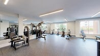 This fitness center is a comprehensively outfitted facility designed to support a full range of workout routines. The space is organized across a generous, multi-zone floor plan that accommodates strength training, cardiovascular exercise, and free weight training simultaneously without crowding. Durable speckled rubber flooring covers the entire area, providing both safety and comfort underfoot throughout each zone.

The strength training area houses a multi-functional cable and smith machine system along with several selectorized weight machines, catering to a wide variety of resistance exercises. A dedicated free weight section features a full rack of dumbbells alongside adjustable benches, offering flexibility for both beginners and advanced users. The cardio zone is equipped with stationary bikes, a rowing machine, and a stair-style climber, positioned near large windows that provide natural light and views of the surrounding outdoor greenery and terrace plantings.

Mirrored walls run along multiple sections of the facility, visually expanding the space and assisting users with form during workouts. Recessed linear LED lighting runs across the ceiling in clean lines, ensuring bright, even illumination throughout. Lockers are conveniently available near the entrance. The overall aesthetic is clean, modern, and neutral, creating a focused and professional training environment.