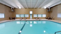 This single-level indoor pool facility offers a welcoming and functional aquatic environment designed for both exercise and leisure. The centerpiece of the space is a generously sized rectangular swimming pool with crystal-clear turquoise water, complete with a designated lap lane for fitness swimming. The pool is surrounded by a smooth, light-toned pool deck that provides ample space for movement and safety. A row of lounge chairs lines the far wall, offering a comfortable area for relaxation before or after a swim. The interior walls feature a warm tan finish, accented by teal-trimmed windows that span the length of the room, allowing natural light to filter in and provide views of the surrounding outdoor environment. A centrally located door with a vertical blind offers direct access to the exterior. The ceiling is finished with a classic white beam structure, adding architectural character while housing overhead lighting that keeps the space well-illuminated throughout the day. The overall ambiance is calm and resort-like, making this indoor pool facility a practical and inviting destination for year-round aquatic activity.