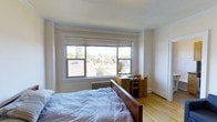 This single-floor studio offers a thoughtfully arranged living space with one bathroom, making smart use of every square foot. The main living area features warm hardwood flooring that lends a clean, inviting aesthetic throughout. A large, multi-pane window serves as a standout architectural element, flooding the room with abundant natural light while framing pleasant views of the surrounding residential neighborhood, complete with mature trees and rooftops. Classic white crown molding accents the ceiling, adding a touch of traditional character to the otherwise neutral, understated palette of soft white walls. A cast-iron radiator beneath the window speaks to the classic construction of the building, contributing to its timeless charm. The layout accommodates a sleeping area and workspace within a compact but functional footprint, with built-in closet storage offering practical organization. The bathroom rounds out the unit, providing all essential amenities. The overall ambiance is calm and airy, with the interplay of natural light and light-toned finishes creating an open, comfortable atmosphere well-suited for everyday living.