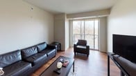 This single-floor residence offers a cohesive and functional layout designed for comfortable everyday living. The kitchen is outfitted with rich dark espresso cabinetry, granite countertops, and a full suite of stainless steel appliances, including a refrigerator, range, microwave, and dishwasher, all set above warm hardwood-style flooring that flows throughout the unit. The living area is open and inviting, featuring large sliding glass doors that lead to a private balcony with views of mature surrounding trees, filling the space with generous natural light. The primary bedroom is generously sized, benefiting from a wall of windows dressed with roller shades and offering peaceful outdoor views. A second bedroom is equipped with a bunk bed setup and a small work surface, making it a versatile space. Both bathrooms are finished with the same granite countertops and dark vanity cabinetry as the kitchen, each offering a tub-shower combination and tiled flooring. The unit maintains a clean, neutral color palette with cream walls throughout, creating a bright and understated ambiance that complements the modern finishes found in every room.