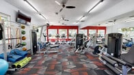 This well-equipped fitness center in Gainesville, Florida offers a comprehensive single-floor workout facility designed to support a wide range of exercise routines. The space is defined by bold red accent walls paired with crisp white ceilings, creating an energetic and motivating atmosphere. Large windows line the walls, flooding the room with natural light and offering views of the surrounding greenery, while ceiling fans contribute to a comfortable workout environment.

The cardio zone features a row of treadmills, elliptical machines, stationary bikes, and a rowing machine, all positioned to take advantage of the natural light and outdoor views. The strength training area is outfitted with a variety of selectorized weight machines and cable systems, along with a dedicated dumbbell rack offering multiple weight options. Full-length mirrors span an entire wall, enhancing the sense of space and allowing users to monitor their form throughout their workout.

A functional training corner houses stability balls in multiple sizes, resistance bands, medicine balls, and exercise mats — providing versatile options for flexibility, core work, and group exercise. A wall-mounted television adds to the amenity. The floor throughout is covered in a durable, patterned carpet in complementary red, gray, and charcoal tones, tying the room's bold aesthetic together cohesively.