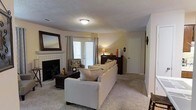 This single-floor residence offers a warm and inviting atmosphere defined by thoughtful design and comfortable living spaces. The living room serves as the heart of the home, centered around a classic tile-surround fireplace with a sleek mounted television, making it an ideal gathering spot. A large, plush sectional sofa in neutral tones anchors the space, complemented by a dark wood coffee table and coordinating furniture pieces that add a grounded, traditional character. The room is finished with wall-to-wall carpeting that enhances the sense of warmth underfoot. Natural light fills the space through a set of elegant French doors dressed with patterned drapes, offering a view and access to the outdoors. A decorative mirror above the fireplace adds depth to the room, while accent lighting from table lamps creates a soft, ambient glow. The textured ceiling adds a classic residential touch throughout. A dark wood dresser or credenza near the French doors provides additional storage and display surface. The overall ambiance is relaxed and homey, with a cohesive neutral color palette of beige and cream tones that create a welcoming environment throughout the space.