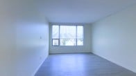 This single-floor space presents a clean, modern aesthetic defined by its minimalist design and bright, airy atmosphere. The interior is characterized by smooth white walls and ceilings that amplify the sense of openness, while light-toned hardwood flooring runs throughout, lending warmth and continuity to the space.

A standout feature is the expansive multi-pane window arrangement, which spans a significant portion of one wall and is fitted with pleated cellular shades. This window configuration floods the room with abundant natural light and offers views of the surrounding residential neighborhood, including mature trees that provide a pleasant, verdant backdrop.

The room's proportions feel generous and well-balanced, with clean wall surfaces that offer versatile potential for various furniture arrangements and uses. Crisp white baseboards frame the perimeter of the room, complementing the overall bright and polished finish of the space. The uncluttered design and neutral palette create a calm, welcoming ambiance that is both functional and visually appealing — an ideal canvas for a range of interior styles.