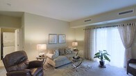 This single-story space showcases a living room designed with both comfort and functionality in mind. The room is finished with wall-to-wall carpeting that adds warmth underfoot, complemented by neutral, beige-toned walls that create a calm and inviting ambiance throughout. A centrally positioned sofa, dressed with decorative pillows, anchors the seating area alongside a sleek glass-top coffee table with a chrome frame, adding a contemporary touch. A leather accent chair rounds out the seating arrangement. Two framed artworks hang symmetrically above the sofa, contributing a polished, coordinated aesthetic. Ambient lighting is provided by matching table lamps on either side of the space, casting a warm glow. A large window dressed with sheer, floor-length curtains allows natural light to filter in softly, brightening the interior. A central air vent is integrated into the ceiling, ensuring climate control throughout. A potted tropical plant near the window introduces a natural element to the decor. The overall atmosphere is well-maintained and move-in ready, offering a functional and tastefully appointed living environment.