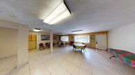 Located at 2630 W Lincoln Ave in Anaheim, California, this single-story community center offers a functional and welcoming space designed to serve a variety of group activities and events. The main gathering hall is the heart of the facility, featuring light-toned tile flooring throughout, warm yellow-painted walls, and overhead fluorescent lighting that illuminates the space evenly. Large windows fitted with vertical blinds line the exterior walls, allowing natural light to filter in and providing views of the surrounding outdoor area. The hall comfortably accommodates round and rectangular tables with seating, making it well-suited for meetings, events, and social gatherings. Structural columns punctuate the open floor plan, adding architectural character while maintaining an expansive feel. A dedicated lounge area features a dark sofa and seating for more relaxed interaction. Adjacent to the main hall is a service counter area with wood cabinetry and a granite-style surface, providing a convenient space for food and beverage setups. A fitness corner is equipped with exercise equipment, adding a wellness dimension to the facility. Security is supported by ceiling-mounted cameras. The center also includes a restroom with tiled flooring and essential fixtures. Overall, the space is practical, accessible, and versatile for community programming.