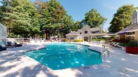 Located at 3409 Mill Tree Rd in Raleigh, NC, this single-level community amenity space is designed to offer a welcoming outdoor retreat. The centerpiece is a generously sized, geometric outdoor swimming pool with crystal-clear blue water, surrounded by a broad, light-colored pool deck outfitted with multiple lounge chairs and umbrellas — ideal for relaxing on sunny days. The pool area is framed by a lush canopy of mature trees that provide natural shade and a serene, park-like atmosphere.

Adjacent to the pool is a covered deck area featuring composite wood decking and a distinctive glass block privacy wall that allows natural light to filter through while providing separation from the surrounding landscape. The covered lounge space is furnished with wicker seating and blue cushioned chairs and sofas, creating a comfortable outdoor living environment. Large open windows in the covered area offer direct sightlines to the pool and the surrounding wooded grounds, enhancing the connection between indoor and outdoor spaces.

The architectural style of the clubhouse building is traditional, with gray lap siding, arched rooflines, and well-maintained landscaping. The overall ambiance of the space is relaxed and resort-inspired, making it a standout communal feature for residents of this Raleigh community.