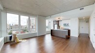 This contemporary single-floor residence offers an open-concept layout that seamlessly blends the living and kitchen areas into one cohesive, light-filled space. The rich, warm-toned hardwood flooring extends throughout, grounding the interior with a sense of depth and continuity. Above, exposed concrete panel ceilings lend an industrial-modern character that contrasts beautifully with the clean white walls and crisp finishes throughout.

The kitchen is a standout feature, anchored by a sleek island finished in dark wood tones with a light countertop surface. White upper cabinetry pairs with darker lower units to create a balanced, modern aesthetic, while stainless steel appliances — including a refrigerator and microwave — provide a polished, functional finish. An elegant multi-bulb chandelier suspended above the island adds a refined focal point to the space.

The living area is generously proportioned and bathed in natural light, courtesy of large floor-to-ceiling windows that frame impressive views of the surrounding urban cityscape. The expansive glazing brings the energy of the city indoors while maintaining a bright, airy atmosphere. Together, these elements create a sophisticated and functional urban living space with a distinctly contemporary character.