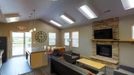 This single-floor space is designed as a welcoming communal area that balances comfort with functionality. The vaulted ceiling is a defining architectural feature, fitted with a row of skylights that flood the interior with abundant natural light, creating an open and airy atmosphere throughout the day. Warm taupe walls and wood-trimmed windows complement the overall neutral palette, lending the space a relaxed yet polished feel.

At the heart of the room sits a stacked-stone fireplace with a built-in wooden mantle, paired with a wall-mounted flat-screen television — forming a natural focal point for the lounge area. Comfortable gray upholstered seating is arranged around a yellow accent coffee table, encouraging relaxed conversation or casual viewing. A large decorative wall clock adds a bold visual element near the transition to the dining zone.

The dining area features a round table with mixed seating, positioned near natural-light windows that offer pleasant outdoor views. Adjacent to this is a kitchenette with a countertop bar, pendant lighting, and cabinetry, supporting light food and beverage needs. Patterned carpeting ties all zones together cohesively.