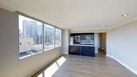 This single-floor residence offers an open-concept living and kitchen area bathed in natural light from large floor-to-ceiling windows that frame striking high-rise city skyline views. The expansive main living space features light-toned hardwood flooring throughout, smooth white ceilings with recessed lighting, and clean neutral walls that create a bright, airy atmosphere.

The kitchen is centrally positioned as a functional island-style unit, finished with deep navy cabinetry on both upper and lower sections, complemented by a mosaic tile backsplash and light stone countertops. Stainless steel appliances, including a built-in microwave and range, add a contemporary touch. The kitchen layout connects seamlessly to the living area, making it well-suited for both everyday living and entertaining.

A hallway visible beyond the kitchen suggests additional rooms further into the unit. The overall design aesthetic is modern and understated, combining dark kitchen accents against light flooring and walls. The prominence of natural light and panoramic urban views from the elevated vantage point are defining characteristics of this space.