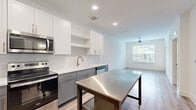 Located at 1290 Old Peachtree Rd NW in Duluth, Georgia, this single-floor residence offers a clean, modern aesthetic throughout. The kitchen serves as a standout feature, showcasing a two-tone cabinet design with crisp white upper cabinets and sophisticated gray lower cabinets, complemented by a classic white subway tile backsplash and bright quartz countertops. A stainless steel island provides additional workspace and storage, while a full suite of stainless steel appliances—including a range, microwave, and dishwasher—rounds out the functional design. Gold-toned hardware and a matching faucet add refined detail throughout. Recessed lighting and large windows bring natural light into both the kitchen and the adjoining living area, which features durable wood-look plank flooring and a ceiling fan. The bedrooms offer plush carpeting, generous window light with views of the surrounding community, and a calm, neutral color palette. A private balcony extends the living space outdoors, overlooking a well-maintained courtyard with mature landscaping and modern architectural surroundings. The overall design balances contemporary finishes with a comfortable, livable atmosphere.