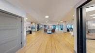Located at 100 Mia Rose Way in Dardenne Prairie, Missouri, this thoughtfully designed single-floor amenity space offers a generous and versatile environment across an expansive open-plan layout. Light oak hardwood flooring runs throughout, complemented by a crisp white drop ceiling with recessed circular lighting that ensures the entire space is bright and welcoming.

The main gathering area serves as the heart of the space, featuring a stylish lounge with leather and upholstered seating arrangements, a linear electric fireplace, a large-screen TV, and a built-in shelving unit — ideal for relaxed socializing or quiet lounging. Adjacent to the lounge is a well-appointed kitchenette with a striking marble-patterned island, navy blue cabinetry, pendant lighting, bar-height seating, and a resident mailbox station along the wall.

Large floor-to-ceiling windows and glass-paneled doors flood the interior with natural light and provide views to the outdoors. Structural columns punctuate the open floor plan, adding architectural rhythm without interrupting the flow. A dedicated fitness center, visible through glass walls, offers equipment for residents. The entrance corridor features sleek black-framed glass partitions and a serene landscape mural accent wall, creating a polished first impression upon arrival.