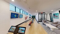Located at 17071 W Dixie Hwy in North Miami Beach, FL, this stylish bowling alley is a standout recreational amenity spanning a generous single-floor layout. The space features two full-length hardwood bowling lanes framed by bold black gutters and crisp white walls, creating a clean, contemporary aesthetic throughout.

A row of clerestory windows runs the length of the lane wall, allowing natural light to filter in while maintaining an energetic, game-ready atmosphere. The sidewall serves as an artistic gallery, displaying a series of eye-catching three-dimensional sculptural installations in teal, navy, and pink, adding a vibrant, creative character to the space. At the far end of the lanes, a lush tropical botanical wallcovering provides a striking focal point.

The bowler's end of the space is thoughtfully appointed with a comfortable lounge area, featuring modern upholstered armchairs and sofas arranged on a bold geometric-patterned area rug. Dual large-screen monitors are mounted above the ball return rack, which holds a full rainbow assortment of bowling balls for players of all skill levels. An adjacent glass-enclosed lounge room offers additional seating for spectators. Recessed ceiling lighting throughout ensures excellent visibility, while the overall design balances function and style seamlessly.