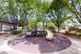 This inviting outdoor recreational area is thoughtfully designed to provide a safe and engaging environment for children and adults alike. At its centerpiece stands a multi-component playground structure finished in green and tan, featuring dual slides, climbing stairs with perforated metal steps, and decorative activity panels. The play area is cushioned by a generous layer of rubber mulch, offering both safety and a clean, well-kept appearance.

Mature deciduous trees are planted throughout the space, forming a lush, shaded canopy that filters natural light and creates a cool, comfortable atmosphere during warmer months. Well-maintained mulched planting beds line the perimeter, filled with ornamental shrubs and groundcover that add greenery and visual interest.

A smooth concrete pathway curves through the courtyard, connecting the playground to a covered seating area that offers a shaded retreat for caregivers and visitors. Classic wood-and-metal benches are positioned along the walkway and near the perimeter fence, providing additional seating. The entire area is enclosed by a sturdy black wrought iron fence, ensuring security while maintaining an open, airy feel. The adjacent building features a combination of brick and modern panel siding, lending an urban yet welcoming character to the overall setting.
