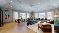 This single-story space is thoughtfully designed with an open-plan layout that seamlessly connects multiple functional areas. The herringbone-patterned hardwood flooring flows throughout, providing a cohesive visual foundation. Vaulted ceilings fitted with multiple ceiling fans and recessed lighting add to the sense of spaciousness and airflow.

The living area is anchored by a striking textured stone accent wall, which houses a wall-mounted television and serves as a bold architectural focal point. A blue sofa, leather armchair, and round coffee table are arranged atop a patterned area rug, creating a comfortable seating arrangement. Colorful abstract artwork adorns the walls, adding vibrancy to the cool grey palette.

Adjacent to the living area, a dining space accommodates a large farmhouse-style wooden table surrounded by mixed seating. Oversized windows with white blinds and linen drapes line multiple walls, flooding the interior with natural light and offering views of the surrounding exterior.

A kitchen area with dark cabinetry, light stone countertops, and an attached bar counter with industrial-style pendant lights and wooden barstools provides both functionality and casual seating. An additional seating and workspace area with café-style tables and chairs occupies the opposite end of the room.