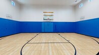 This single-level indoor basketball court is a well-appointed athletic facility designed for serious play and safety. The court features a classic hardwood-style sprung floor finished in a light natural tone, with clearly marked black regulation lines outlining the half-court layout, including a painted arc and key. A wall-mounted glass backboard with a standard hoop is positioned at one end, supported by a wooden mounting bracket secured to the upper wall.

Safety is a clear design priority throughout the space. Vibrant royal blue padded wall panels line the perimeter from floor to mid-wall height, providing full-surround impact protection. The upper portion of the walls transitions to a clean white finish, contributing to an open, bright atmosphere. Recessed ceiling lighting, supplemented by a row of clerestory windows positioned near the roofline, ensures the court is well-lit during all hours of use. The high ceilings provide generous vertical clearance appropriate for gameplay.

Access points are integrated discreetly into the padded wall system, with exit signage clearly marked. HVAC vents are visible along the upper walls, ensuring proper ventilation throughout the enclosed space. The overall design is functional, clean, and purpose-built for athletic use.