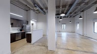 Studio 203 is a single-floor space with a distinctive industrial aesthetic that blends raw architectural elements with functional design. The expansive open floor plan is anchored by polished concrete floors and structural concrete columns, lending the space a loft-like character with a sense of generous, flexible square footage.

The ceiling features exposed ductwork, concrete beams, and pendant lighting — a combination that reinforces the industrial character while providing ample illumination throughout. A window along the far wall allows natural light to filter into the main open area, adding brightness to the otherwise minimalist interior.

A kitchenette is integrated along one wall and includes dark wood cabinetry, a refrigerator, built-in shelving, and a white peninsula that subtly separates the kitchen zone from the larger open area. This layout allows the remainder of the studio to serve as a versatile, unobstructed space suited for a variety of configurations.

The white-painted walls contrast with the raw ceiling and flooring, creating a clean, modern backdrop. Overall, Studio 203 presents a well-balanced mix of industrial character and functional utility.