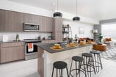This thoughtfully designed single-floor residence showcases a seamless open-concept layout that connects the kitchen, dining, and living areas into one cohesive, flowing space. The kitchen is a standout feature, outfitted with full-height flat-panel cabinetry in a warm wood-tone finish, stainless steel appliances including a refrigerator, range, and microwave, a subway tile backsplash, and a generous island with a dark countertop that provides casual bar-style seating beneath a pair of sleek pendant lights. Light wood-look flooring runs throughout the main living area, enhancing the bright and airy atmosphere. The living room is anchored by a large picture window that frames sweeping panoramic views of the surrounding cityscape and hills, filling the space with abundant natural light. The bedroom continues the tranquil aesthetic with soft carpeting, a large window offering the same scenic views, and warm neutral tones throughout. The bathroom is clean and modern, featuring a wood-framed vanity with a dark stone countertop, an undermount sink, and crisp tile flooring. Throughout the residence, the design balances contemporary finishes with a warm, livable character, creating a comfortable and visually cohesive environment.