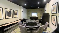 Located in Dallas, TX, this single-floor entertainment space is designed with leisure and socializing in mind. The game room serves as the centerpiece of the space, thoughtfully appointed with a variety of entertainment options to accommodate different tastes and group sizes. A large round poker table surrounded by tufted grey leather chairs commands the center of the room, providing an inviting setting for card games and friendly competition. A full-length shuffleboard table lines one wall, adding a classic recreational element to the space. An upright arcade machine stands nearby, offering a nostalgic gaming experience. The room is anchored by a wall-mounted flat-screen television positioned above a dark wood credenza, making it equally suited for watching sports or movies. Decorative metallic wall sculptures add an artistic touch against the textured accent wall, contributing to the room's modern and sophisticated ambiance. Recessed lighting set against a bold black ceiling creates a moody, lounge-like atmosphere, while wall sconces provide warm, ambient illumination. The space strikes a balance between stylish interior design and functional entertainment, making it an ideal venue for gatherings and recreational activities.