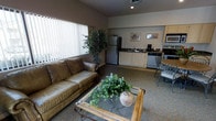 This single-floor space showcases an open-concept layout that seamlessly connects the living area to the kitchen, creating a functional and cohesive environment. The living area is anchored by a brown leather sofa paired with a glass-top coffee table accented by natural greenery, lending a relaxed, earthy ambiance to the space. Neutral-toned carpeting runs throughout, complementing the warm, muted wall colors.

Large vertical blinds on a sizable window allow for adjustable natural light, brightening the main living space. A tall indoor plant beside the window adds a touch of organic décor and enhances the welcoming atmosphere.

The kitchen area is well-equipped with full-sized stainless steel appliances, including a refrigerator, stove, dishwasher, and microwave, alongside light wood cabinetry and a sink with ample counter space. A small dining set with metal chairs is positioned near the kitchen, providing a convenient dining area.

Framed artwork on the kitchen wall adds a decorative touch, while recessed lighting ensures consistent illumination throughout. Overall, the space offers a comfortable, practical layout suited for everyday living.